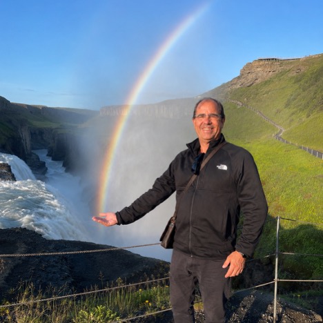 John Waller beside a waterfall and rainbow in Iceland